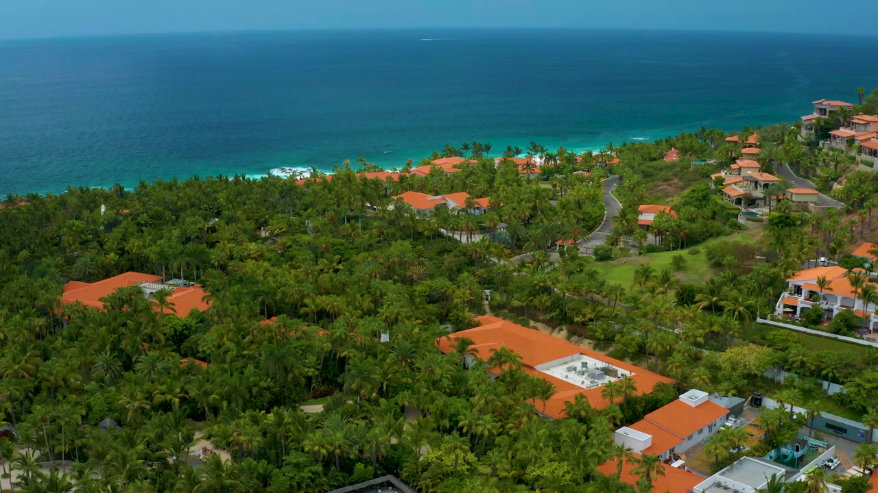Aerial view over Palmilla Beach Cabo, Mexico, with dense green trees, red-roof buildings, a blue sea horizon, and no visible people.