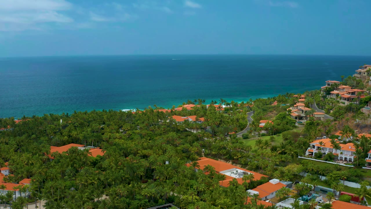 Early frame from Palmilla Beach Cabo, Mexico, showing a level ocean horizon, green palms, orange-roofed buildings, a road through the hillside, and no visible people.