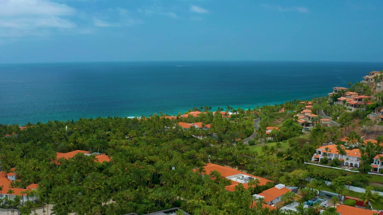 Aerial frame over Palmilla Beach Cabo, Mexico, with more open ocean across the top, a dense green midground, scattered orange rooftops, hillside residences at right, and no visible people.