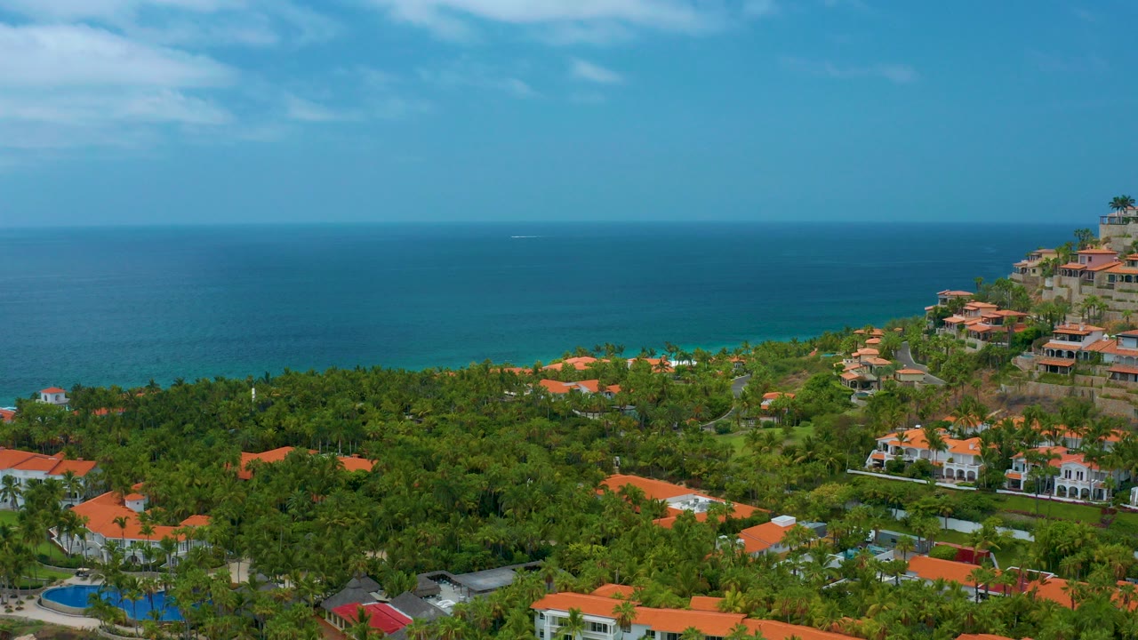 Opening aerial frame at Palmilla Beach Cabo, Mexico, with orange-roofed resort buildings among dense green trees, a hillside at right, blue ocean beyond, and no visible people.