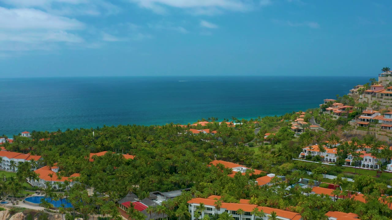 A later aerial frame over Palmilla Beach Cabo shows a wide band of ocean, green resort landscaping, scattered orange roofs, right-side hillside development, and no visible people.