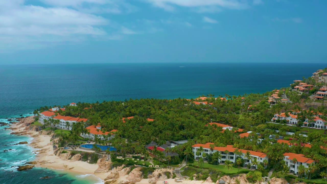 Wide coastal aerial with sandy cove, rocks, palm trees, homes, and open ocean.