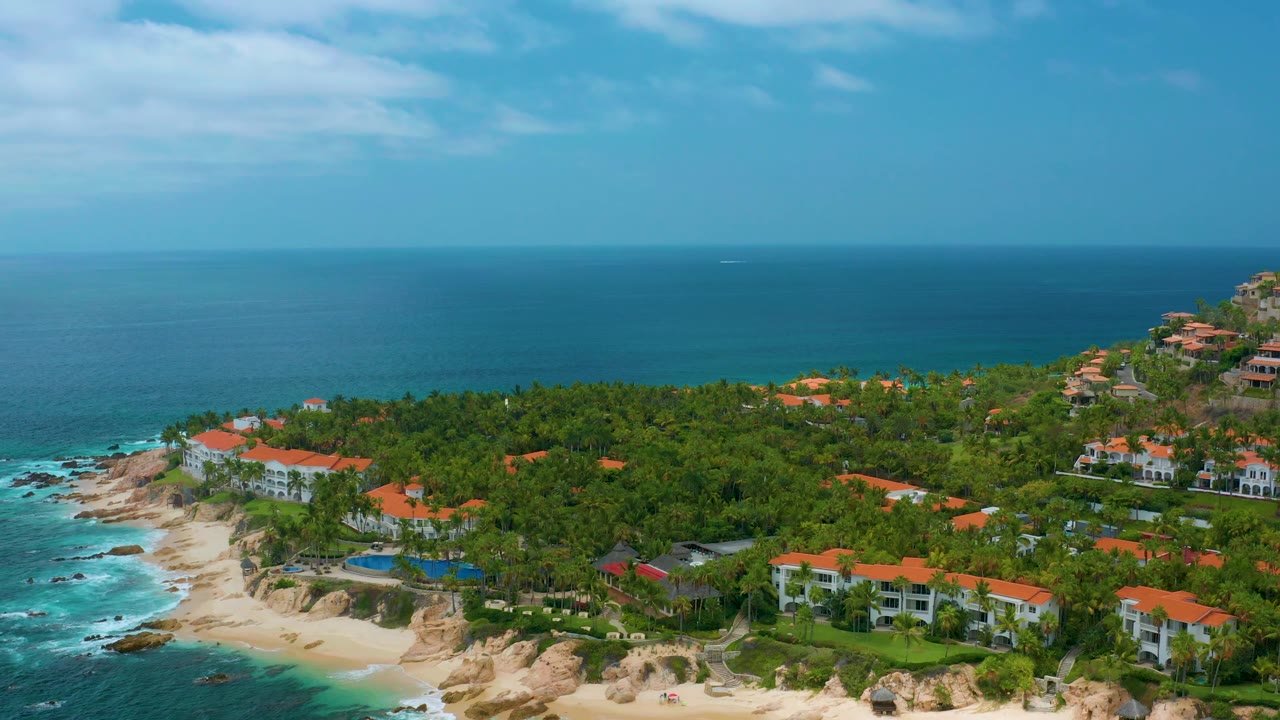 Palmilla Beach Cabo aerial view with surf along the left shoreline, red-roofed homes among palms, hillside buildings to the right, and no visible people.