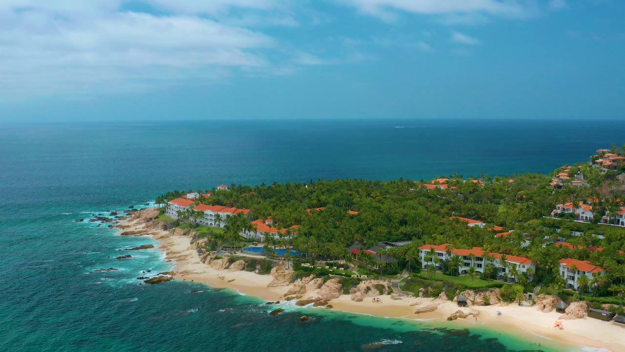 Aerial view over Palmilla Beach Cabo, Mexico, with turquoise water, pale sand, rocky shoreline, palm-covered resort grounds, red-roof buildings, and no visible people.