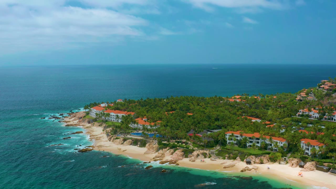 Wide aerial frame at Palmilla Beach Cabo, Mexico, showing surf along the rocky cove, a bright sand strip, palm trees, red-roof resort buildings, open sea, and no visible people.