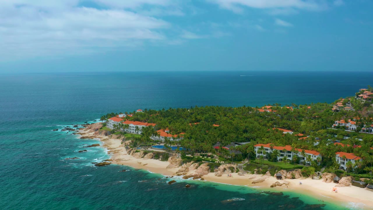 Drone-height view over Palmilla Beach Cabo, Mexico, with teal water in the foreground, waves around rocks, a palm-covered point, red-roof buildings, and no visible people.