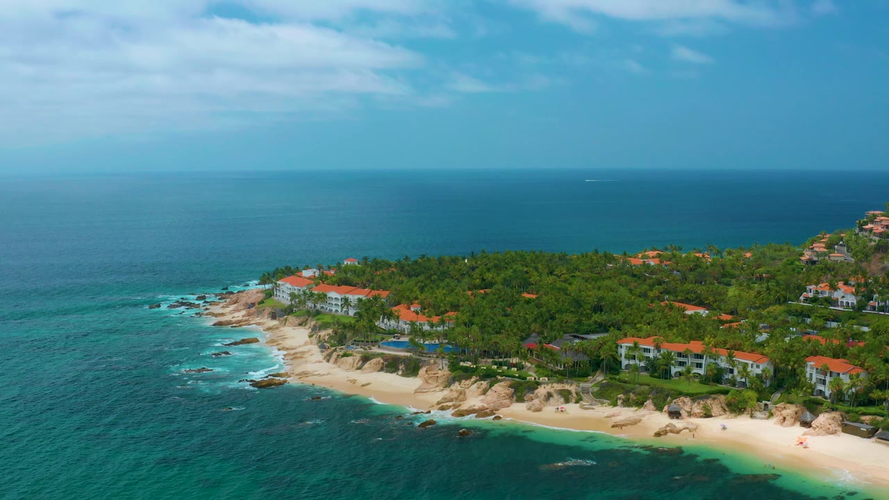 Aerial view of beach cove, resort buildings, palms, and blue horizon.