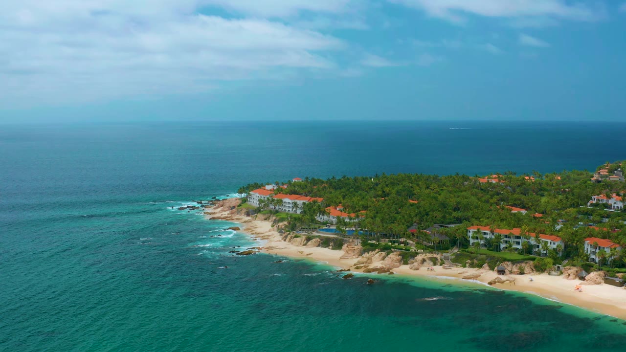 High coastal view with blue water, beach, palms, and red-roofed buildings.