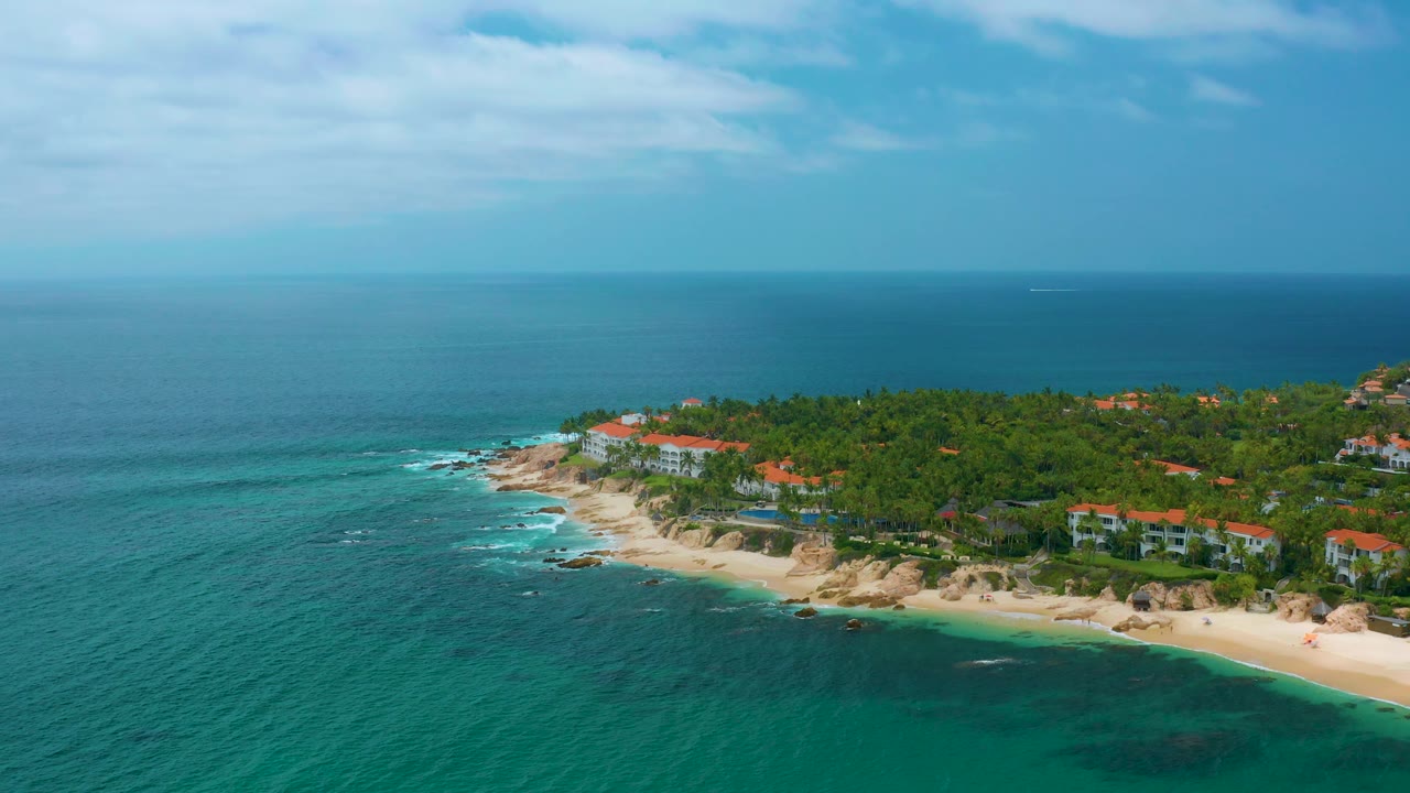 Elevated Palmilla Beach Cabo, Mexico frame with more open water in front, a pale beach curving along rocky points, low coastal buildings behind palms, and no visible people.