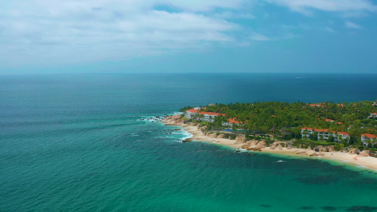 Wide aerial coastline with clear water, beach, palms, and red-roofed houses.