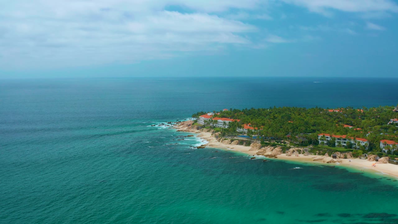 A high aerial view of Palmilla Beach Cabo shows the ocean stretching across most of the frame, with the beach and palm-covered residential shoreline set along the right side.
