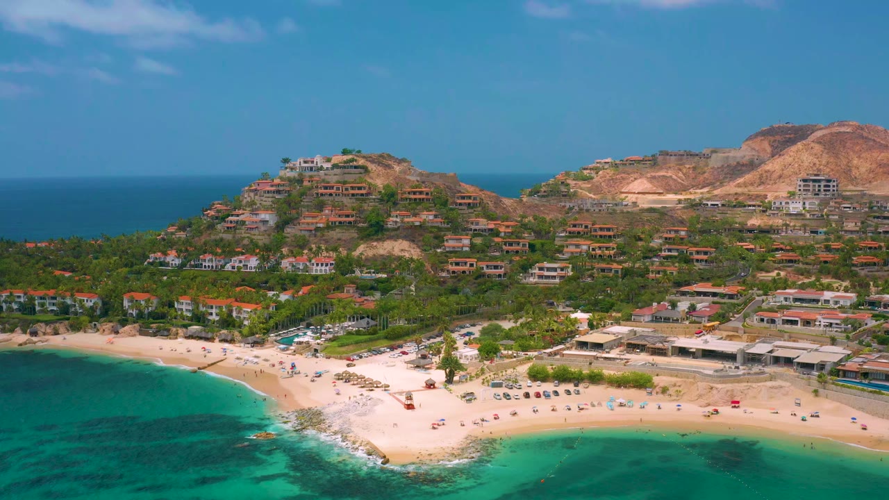 Opening aerial frame over Palmilla Beach Cabo, Mexico, with turquoise sea at lower left, a pale sandy shore, resort buildings, hillside homes, dry mountains, blue sky, and no visible people.