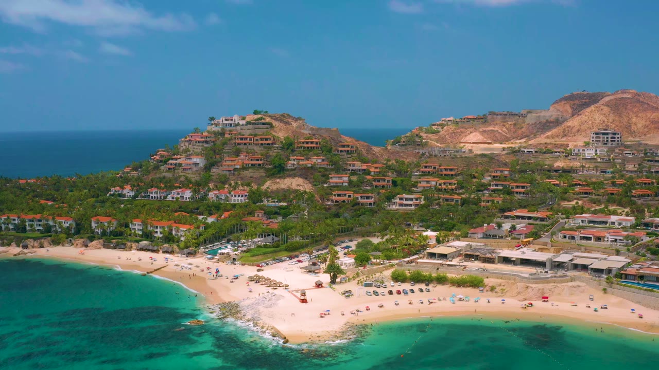 Aerial view at 0:01 over Palmilla Beach Cabo, Mexico, showing more of the curved sandy beach, turquoise water, resort roofs, hillside residences, mountains, blue sky, and no visible people.
