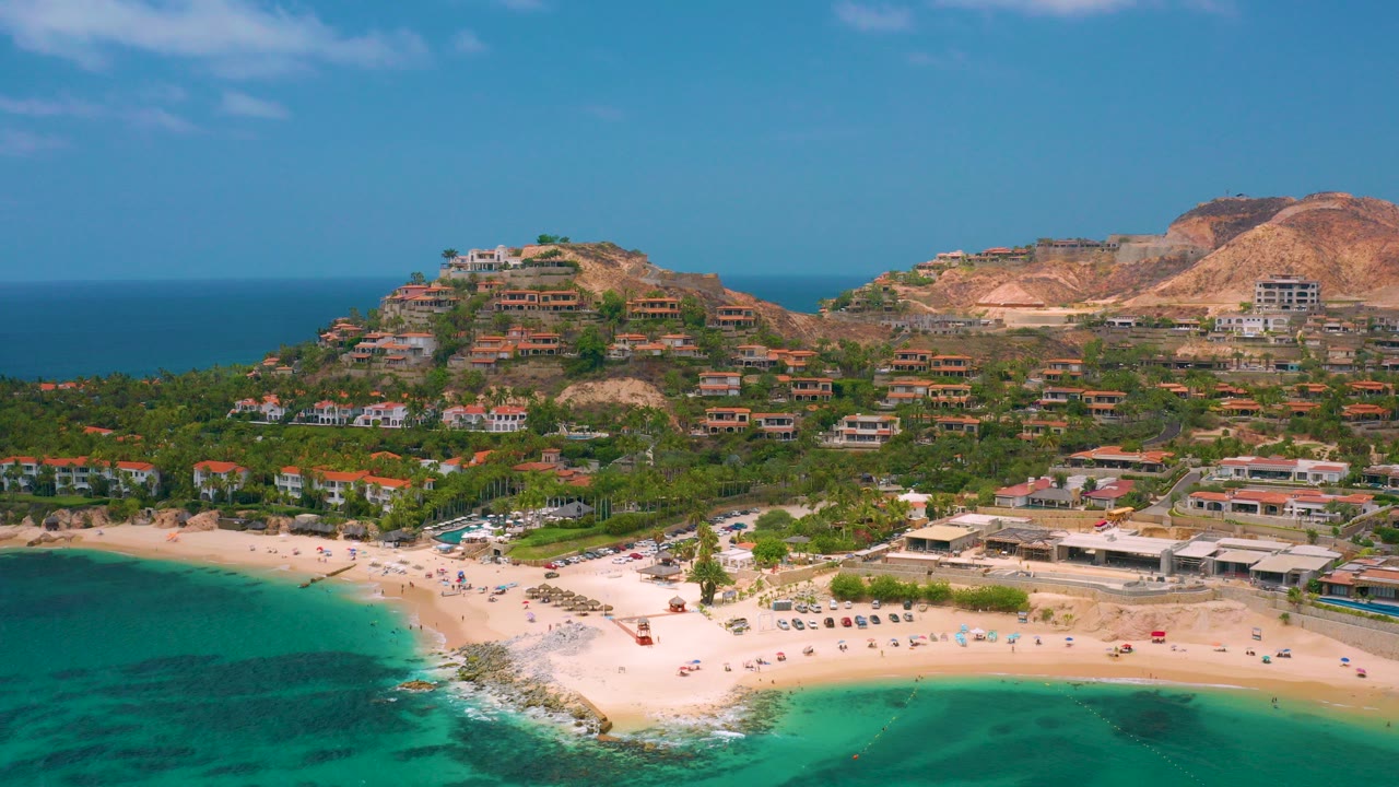 Opening aerial frame over Palmilla Beach Cabo, Mexico, with turquoise water in front, a pale sand curve, hillside homes, dry ridges, blue horizon, and no visible people.
