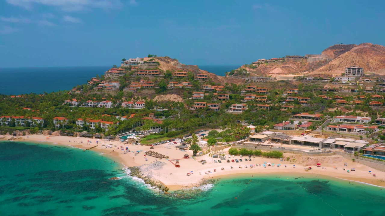 Wide drone view of beach water, shoreline buildings, palms, and hillside houses.