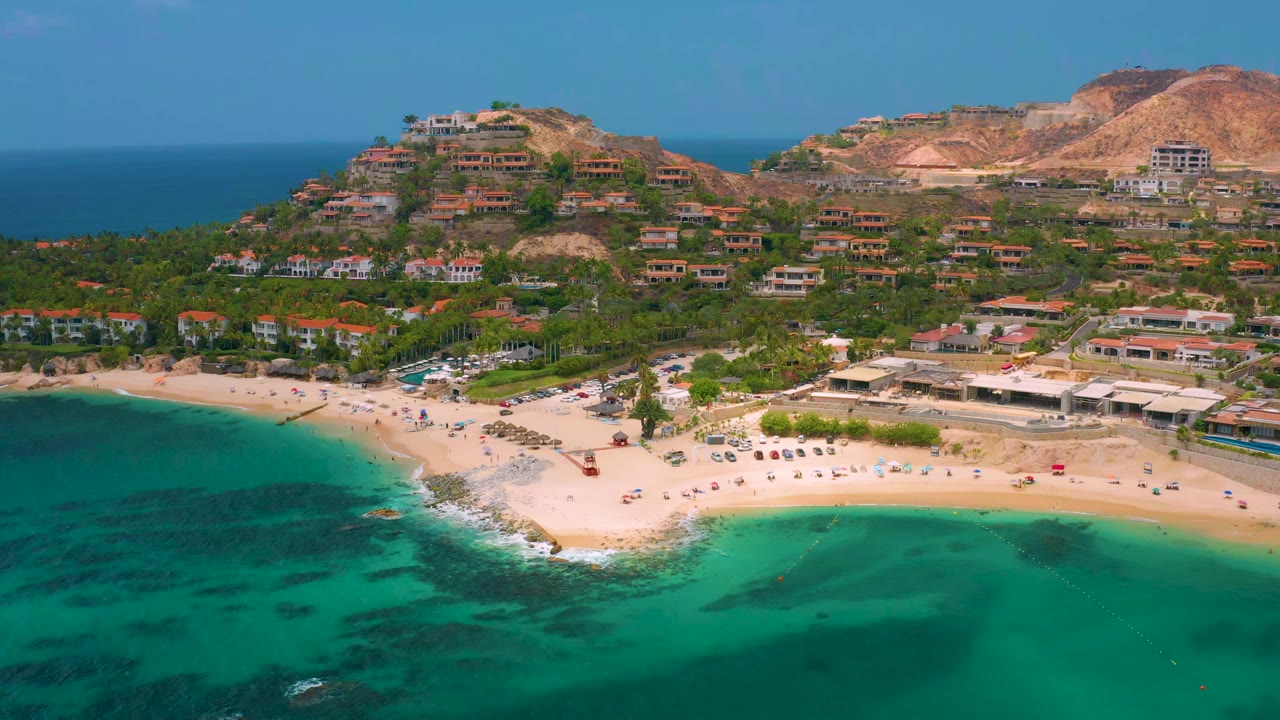 High coastal view with green-blue water, beach umbrellas, vehicles, hillside homes, and mountains.