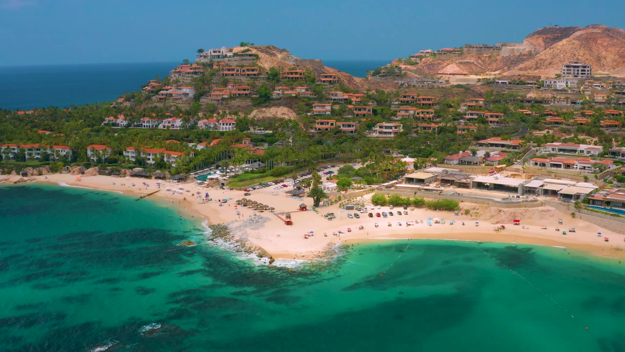 Palmilla Beach Cabo aerial frame with a broad turquoise bay in front, a light sand beach with small shore objects, hillside residences across the background, dry mountains to the right, and no visible people.