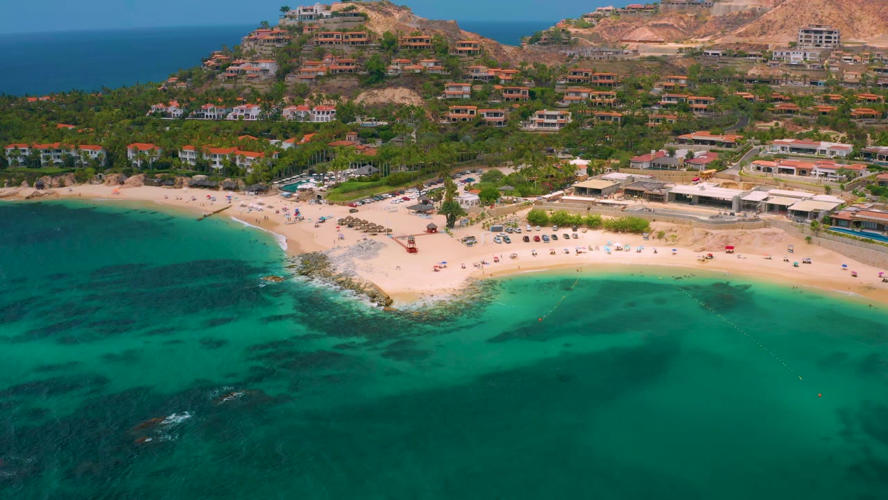 Aerial view of Palmilla Beach Cabo, Mexico, with green-blue water covering the lower frame, a pale beach band in the middle, hillside buildings above, and no visible people.