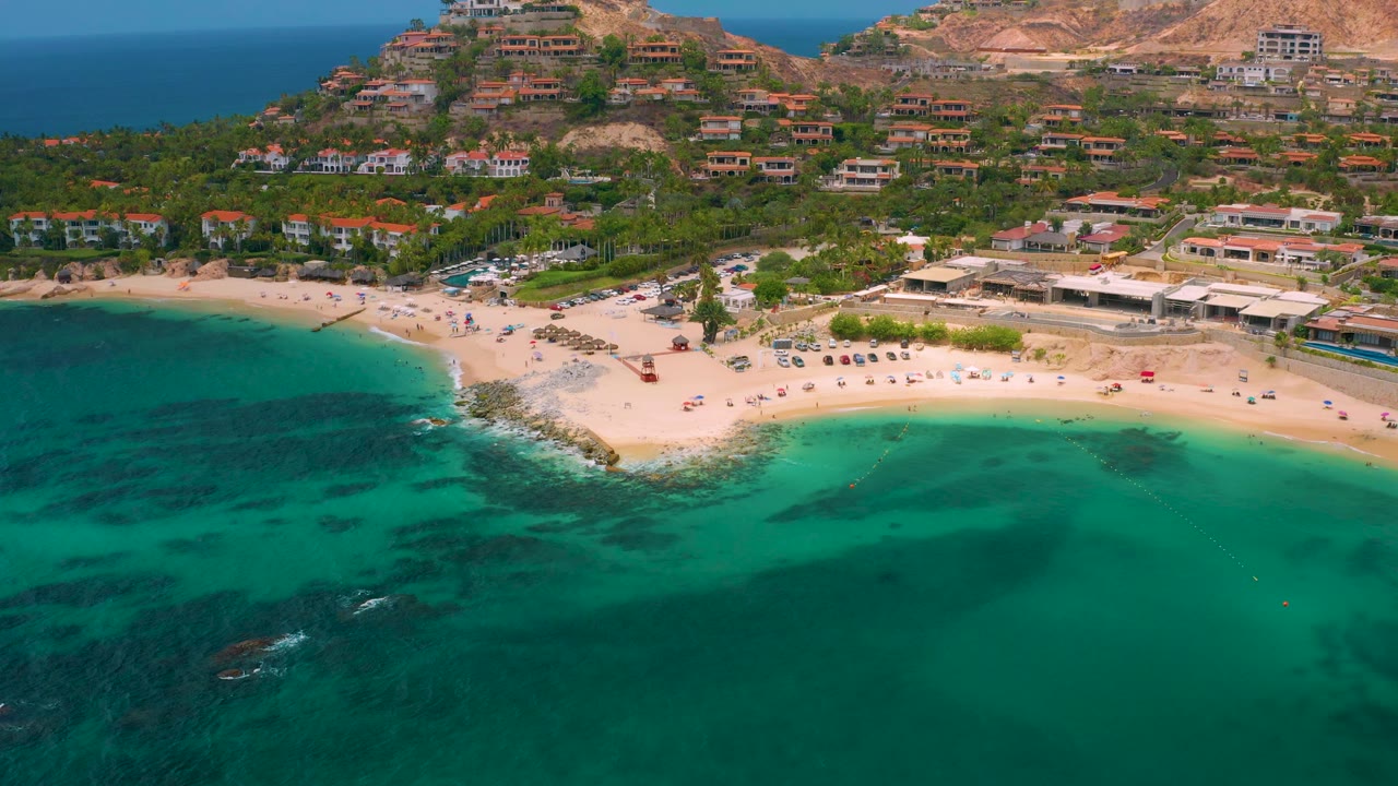 Palmilla Beach Cabo, Mexico from above, showing a broader sweep of turquoise shallows, beach umbrellas along the sand, palm-lined grounds, hillside homes, and no visible people.