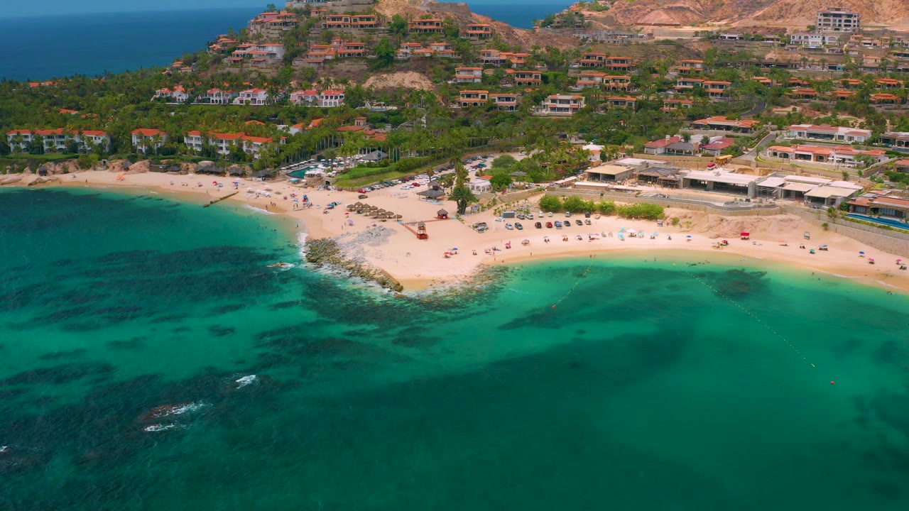 Aerial coastline view with sandy beach, clear water, buildings, and hillside homes.