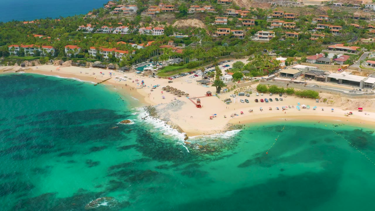 Opening aerial frame over Palmilla Beach Cabo, Mexico, with green water in the foreground, pale sand and surf in the middle, hillside homes beyond, and no visible people discernible.