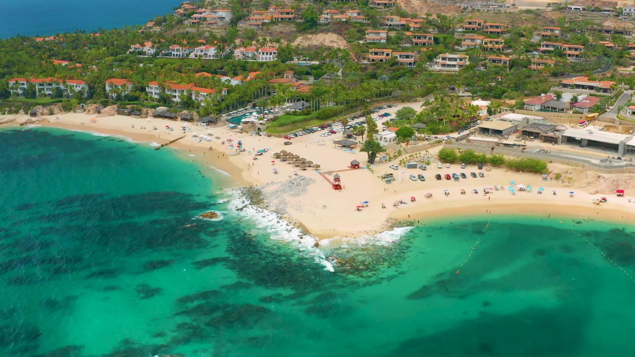 Palmilla Beach Cabo, Mexico from above, with dark underwater patches in green water, surf breaking near rocks, a broad sandy beach, beach structures, hillside homes, and no visible people discernible.