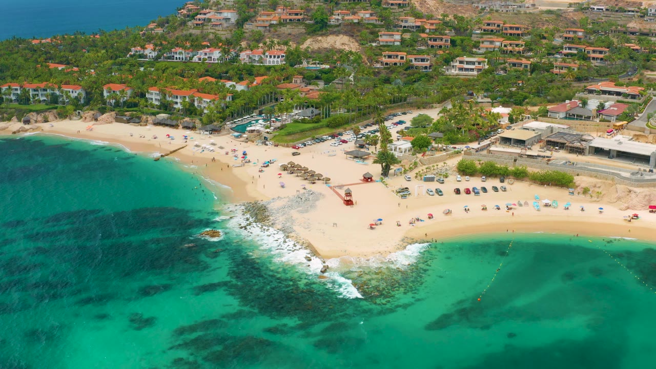 Aerial beach view with surf washing around rocks, green water, parked cars, palms, and homes on hills.