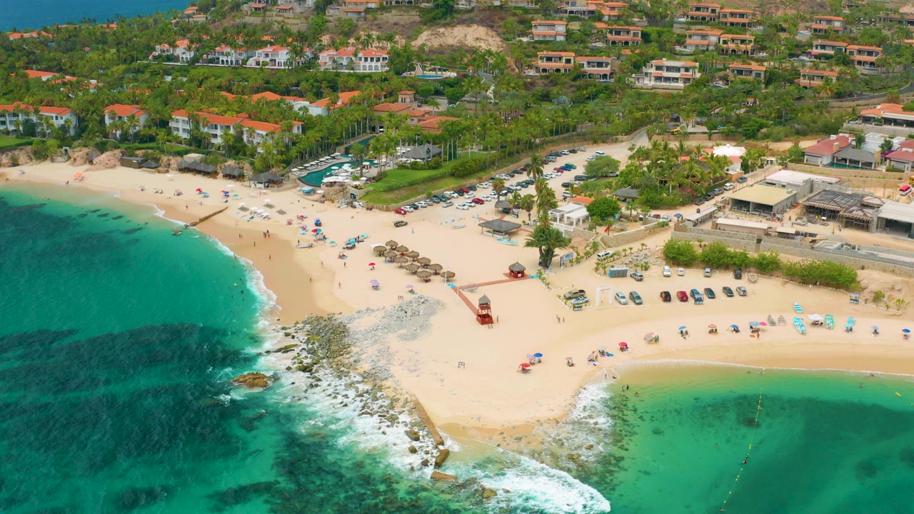 High aerial frame over Palmilla Beach Cabo, Mexico showing teal water on both sides of a sandy beach, umbrellas, parked cars, beach structures, hillside homes, and tiny distant beachgoers.