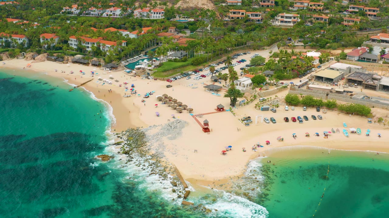 Palmilla Beach Cabo, Mexico appears from above with a broad crescent of sand, darker underwater patches, palapa shades, rows of parked cars, beach buildings, hillside homes, and tiny distant beachgoers.