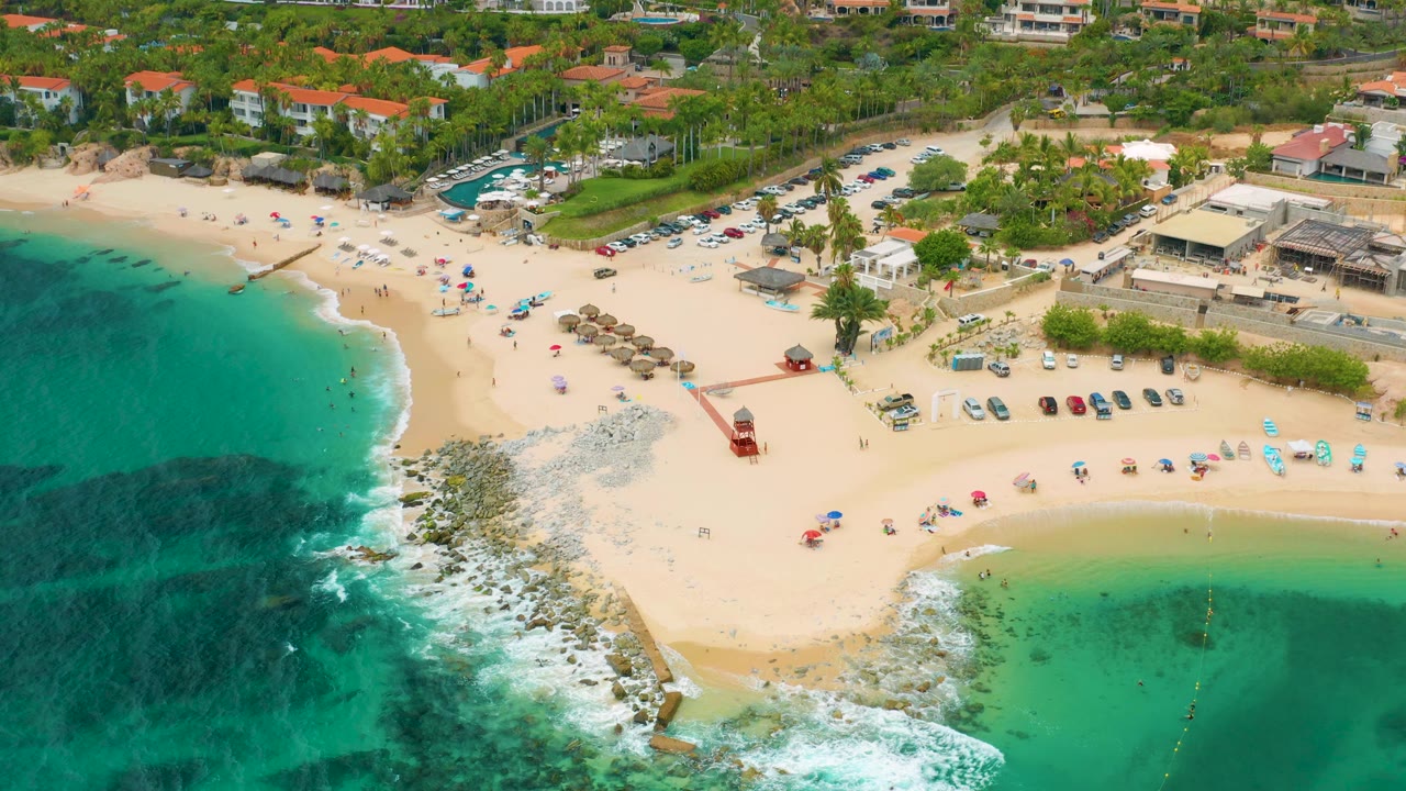 Drone view of beach, rocky point, turquoise surf, umbrellas, and coastal buildings.