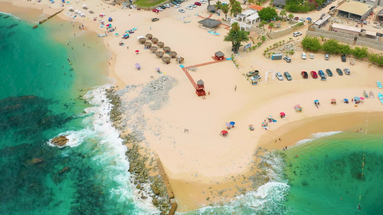 Aerial beach scene with waves breaking on rocks beside umbrellas and parked cars.