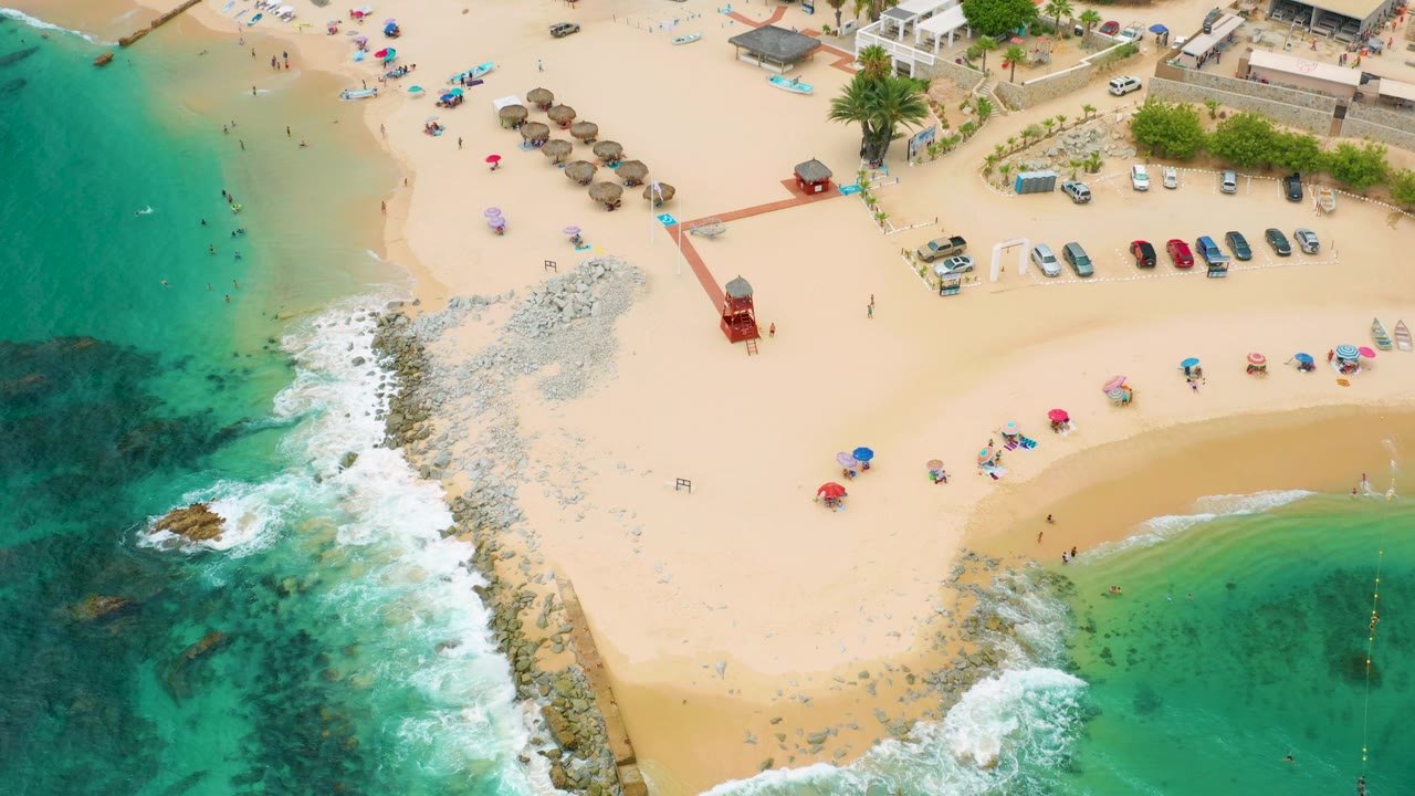 Drone view at Palmilla Beach Cabo, Mexico, with a rocky point between green water and pale sand, a row of parked cars above the beach, and beach-goers spread along the shoreline.