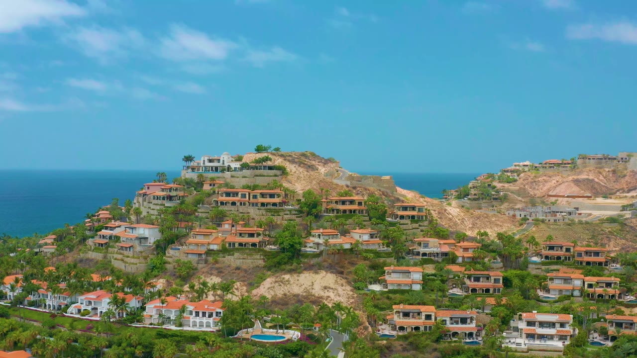 Early frame of Palmilla Beach Cabo hillside homes with orange roofs, palms, exposed earth, blue ocean behind the ridge, and no visible people.