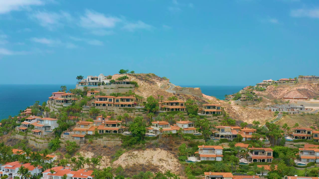 Palmilla Beach Cabo hillside homes fill the center of the frame, with tan graded slopes to the right, blue sea on the horizon, and no visible people.