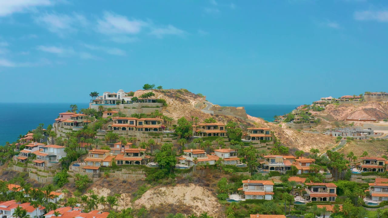 Elevated Palmilla Beach Cabo view with a white hilltop residence, rows of orange-roofed homes, palms, tan cliffs, ocean horizon, and no visible people.