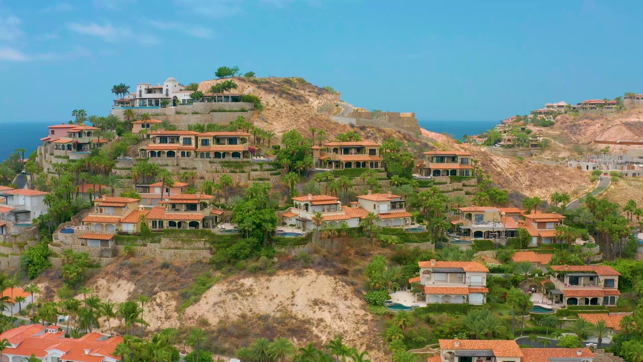 Opening frame of Palmilla Beach Cabo, Mexico hillside homes with terracotta roofs, green landscaping, rocky slopes, a sliver of ocean, and no visible people.