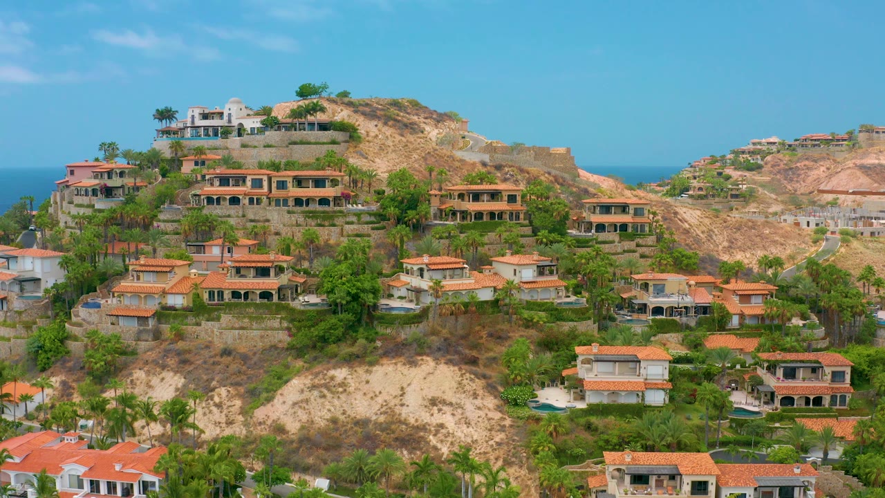 Aerial frame of Palmilla Beach Cabo homes spread across a rocky slope, with palms and shrubs in front, ocean visible behind the ridge, and no visible people.