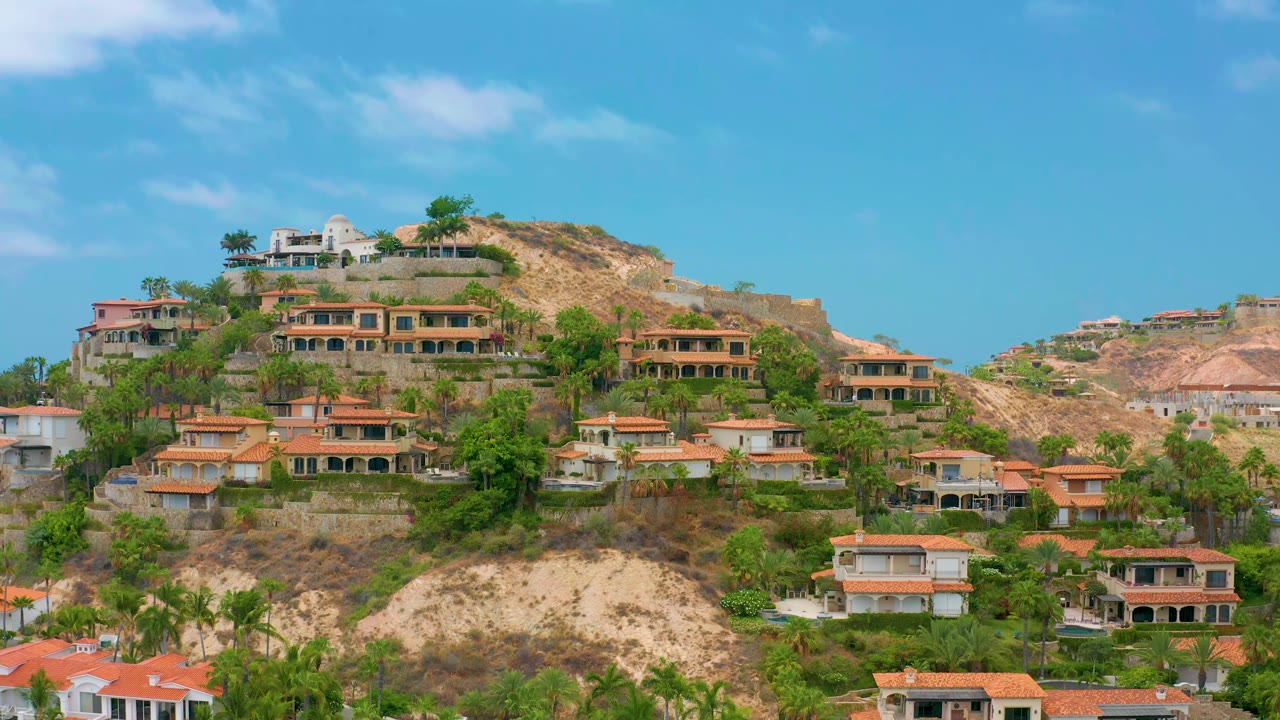 A similar Palmilla Beach Cabo residential hillside view shows layered homes, palms and trees, tan exposed earth, blue sky, and no visible people.