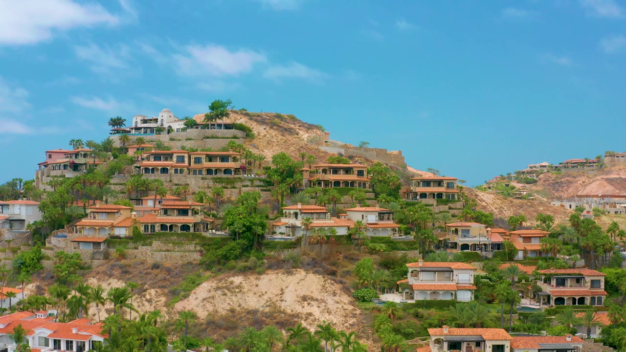 Palmilla Beach Cabo homes line the slope in Mexico, with a white structure near the ridge, orange rooflines below, dry hills, green plantings, and no visible people.