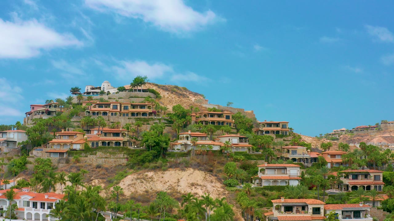 Opening view of Palmilla Beach Cabo hillside homes with tile roofs, palms, dry slope, blue sky, and no visible people.