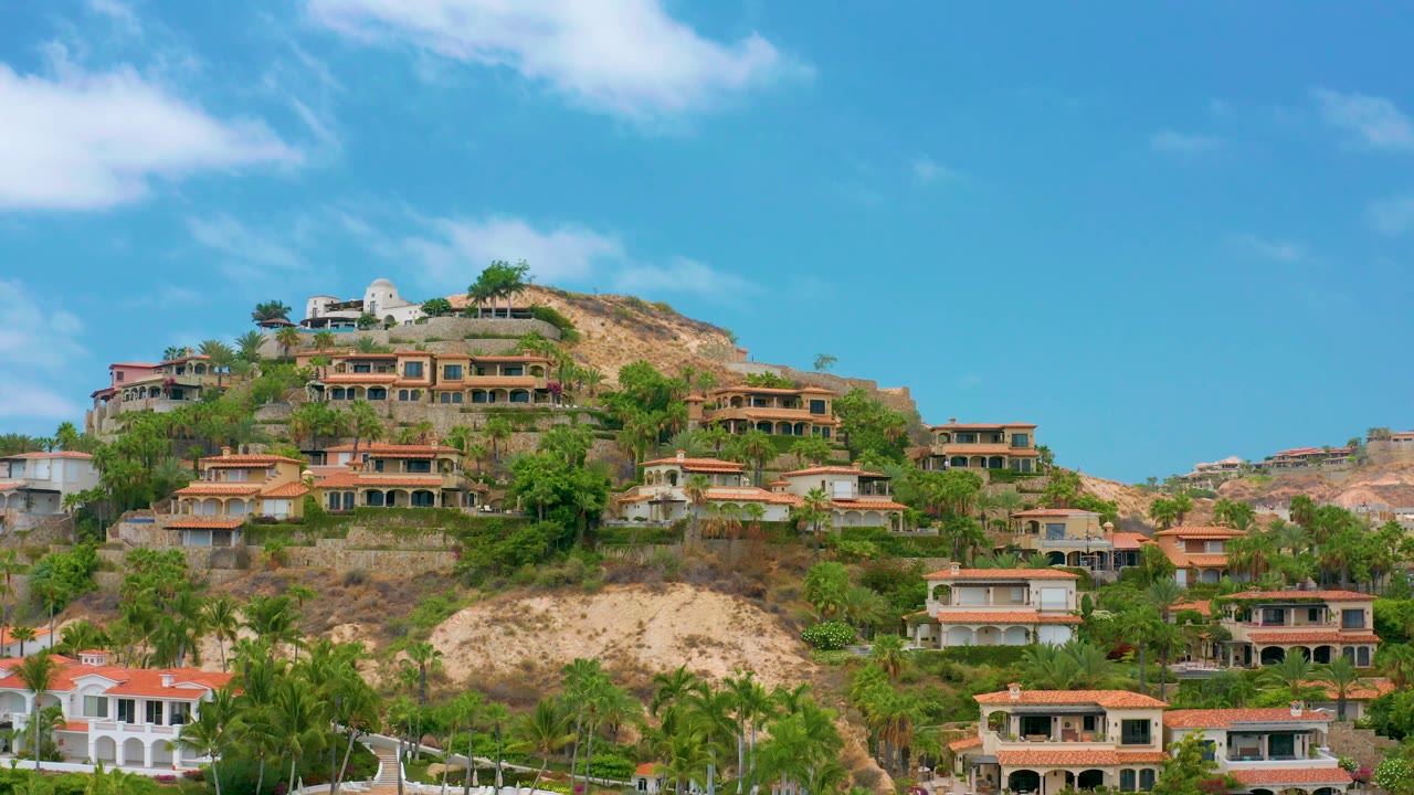 Wide view of Palmilla Beach Cabo hillside buildings, red and tan roofs, green palms, scattered clouds, and no visible people.