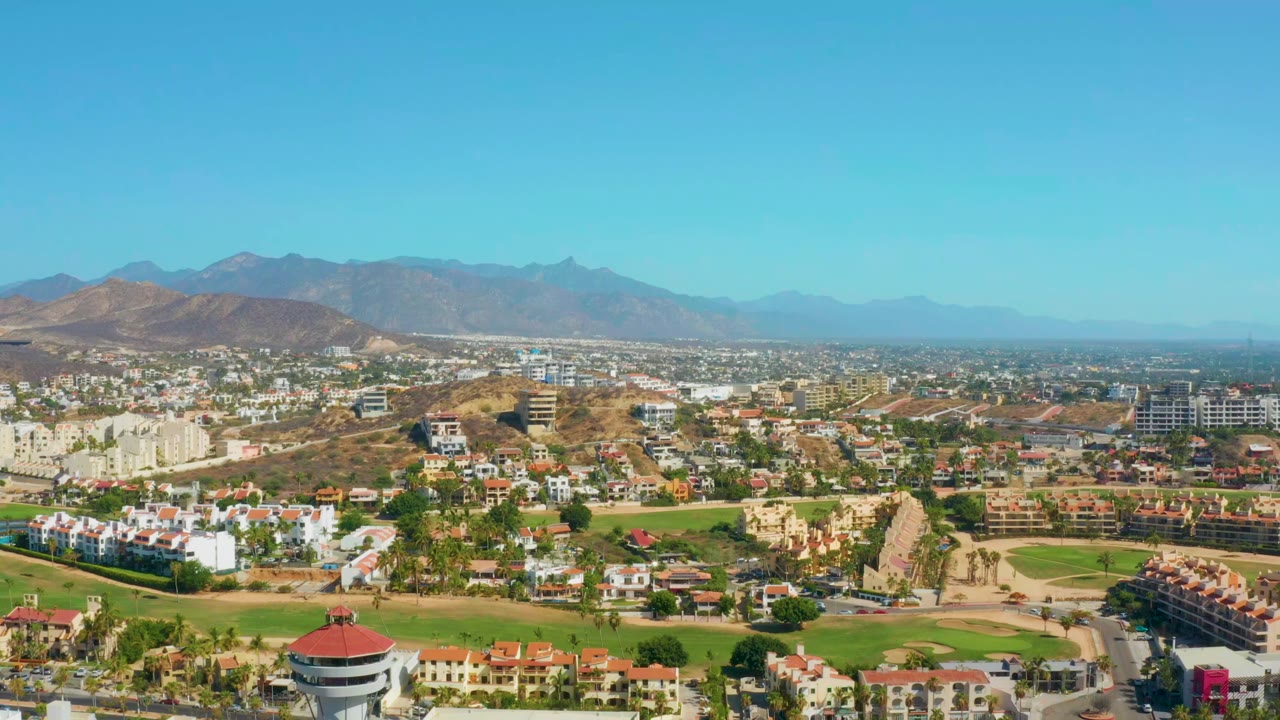 Opening aerial view over San Jose Del Cabo, Mexico, with white buildings, green grounds, dry hills, distant mountains, clear blue sky, and no visible people.