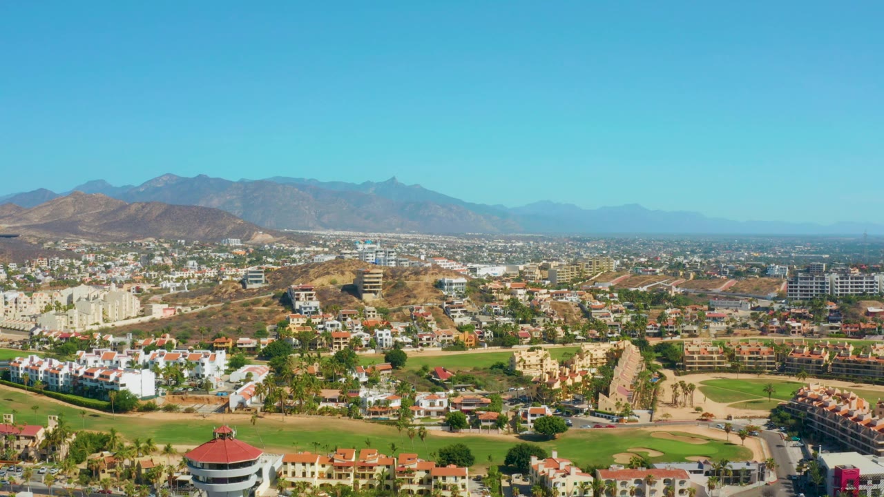 Aerial frame over San Jose Del Cabo with low-rise development spread across the valley, brown hills behind it, mountains on the horizon, and no visible people.