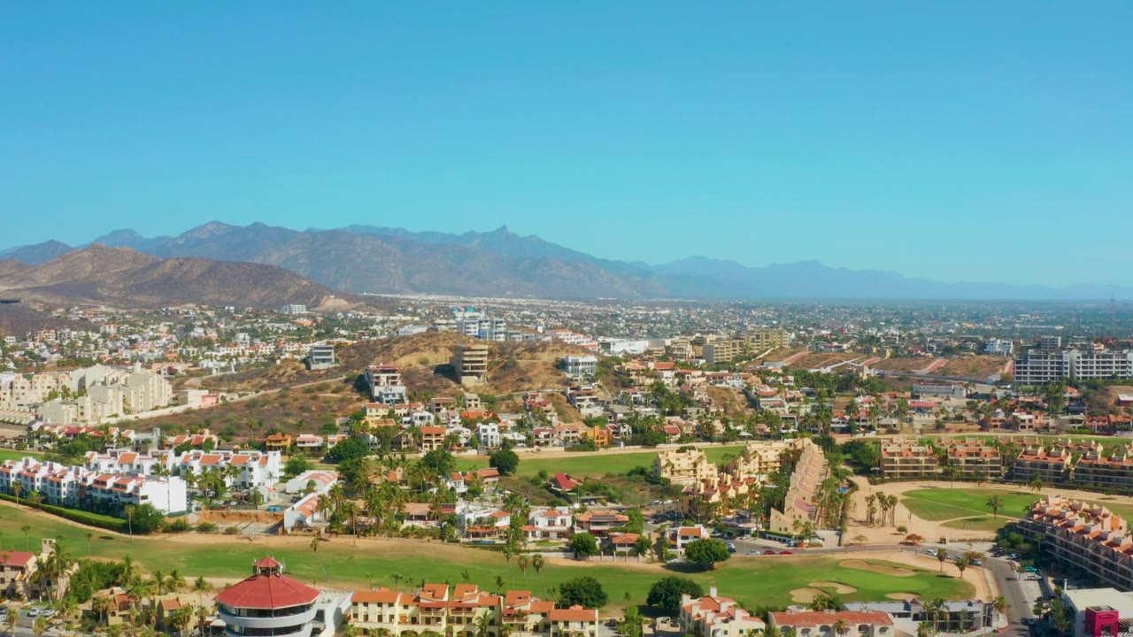 City valley aerial with golf-like green areas and mountain backdrop.