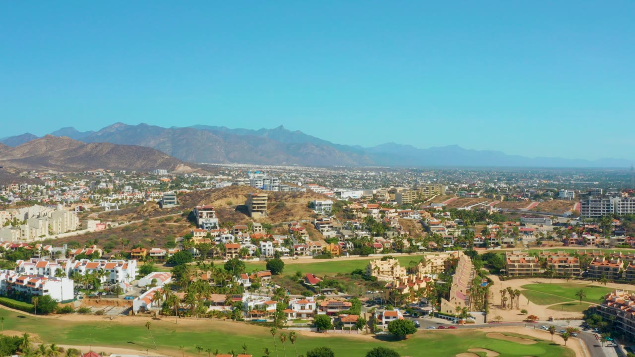 Opening aerial frame over San Jose Del Cabo with golf greens in the foreground, low buildings across the town, mountains in the distance, and no visible people.