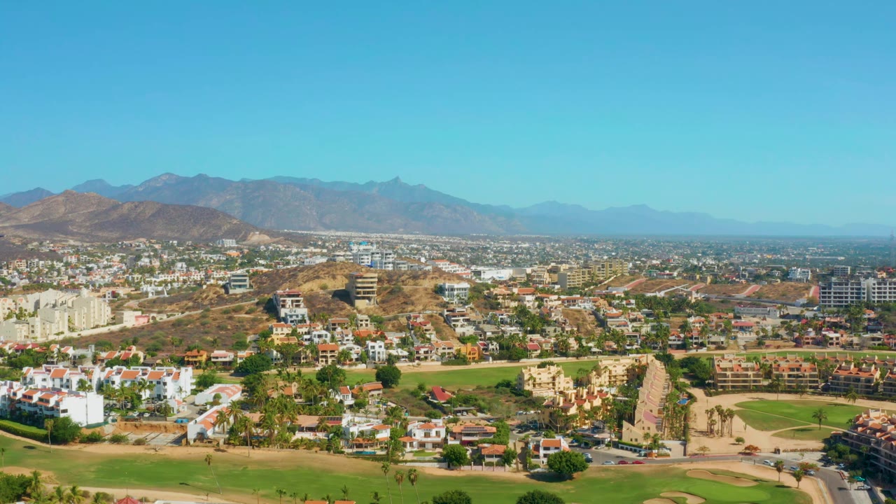 San Jose Del Cabo aerial view one second in, showing white homes, tan resort-style buildings, green fairways, roads, dry hills, distant mountains, and no visible people.