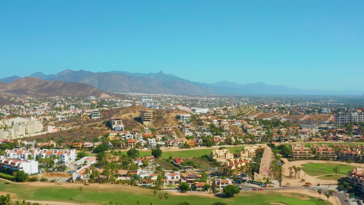 A high daytime view across San Jose Del Cabo with the golf course low in frame, clustered buildings through the middle, hazy mountains beyond, and no visible people.