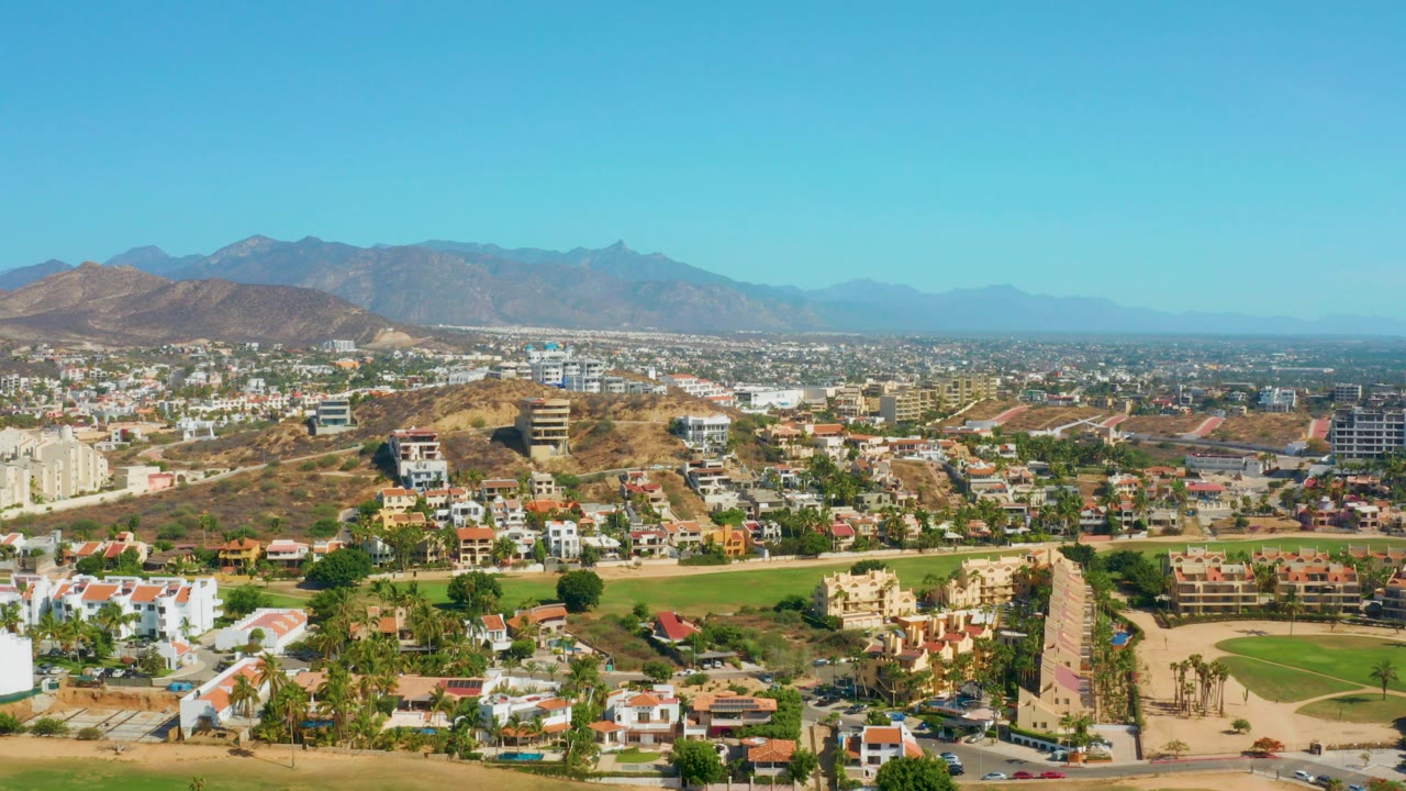 Opening aerial frame over San Jose Del Cabo, Mexico, showing low-rise buildings, a green fairway or park, dry hills, distant mountains, clear blue sky, and no visible people.