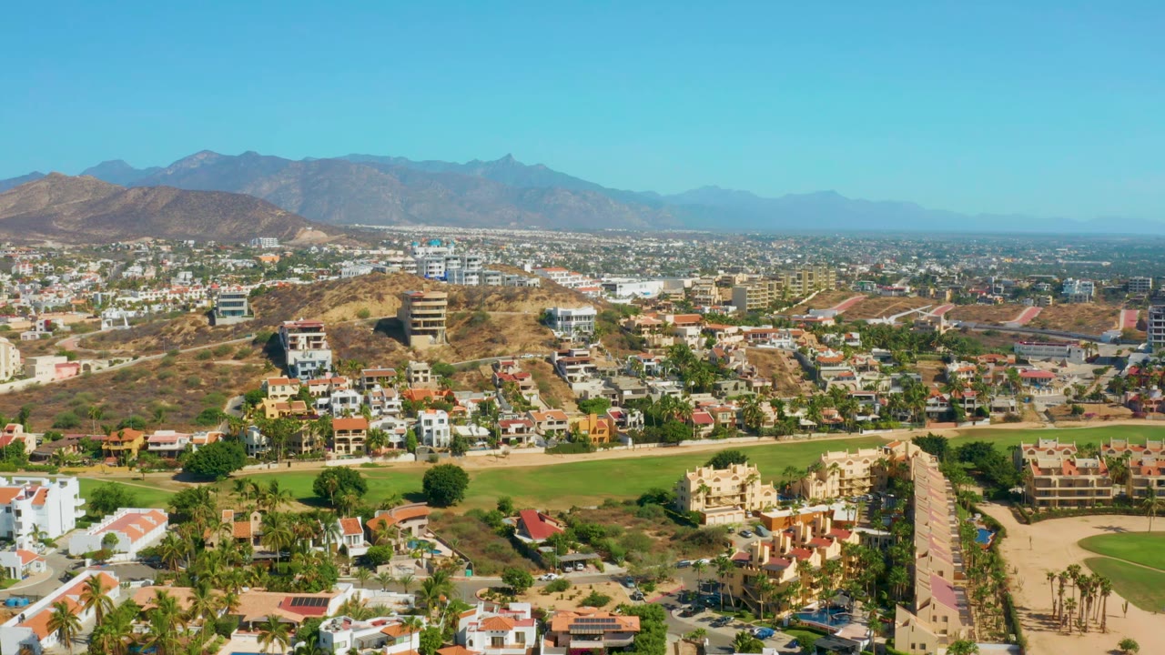 One second into the clip, the San Jose Del Cabo aerial view holds on residential blocks, tan hills, green fairway-like landscaping, mountain horizon, and no visible people.