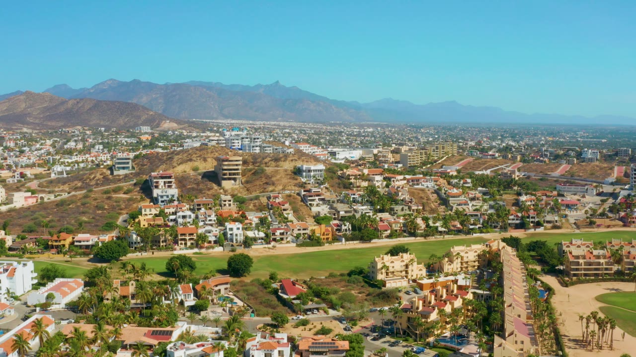 The frame shows San Jose Del Cabo from above with homes and roads in the foreground, open green landscaping across the lower frame, dry hills behind town, and no visible people.